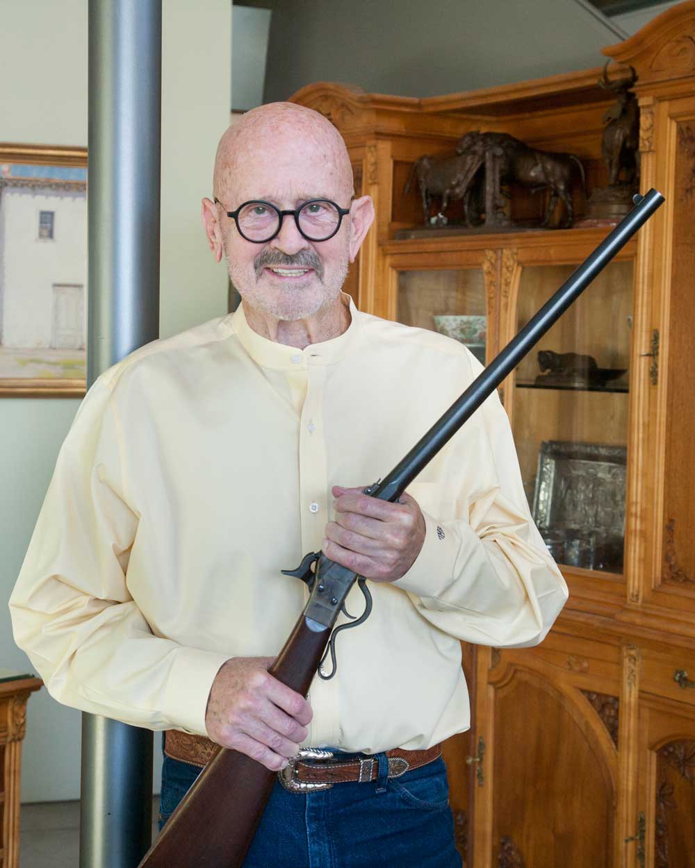 Brad Witherell, an older gentleman, stands facing the camera holding an antique rifle. He is wearing a long-sleeved, yellow button-up shirt, belt, and jeans.