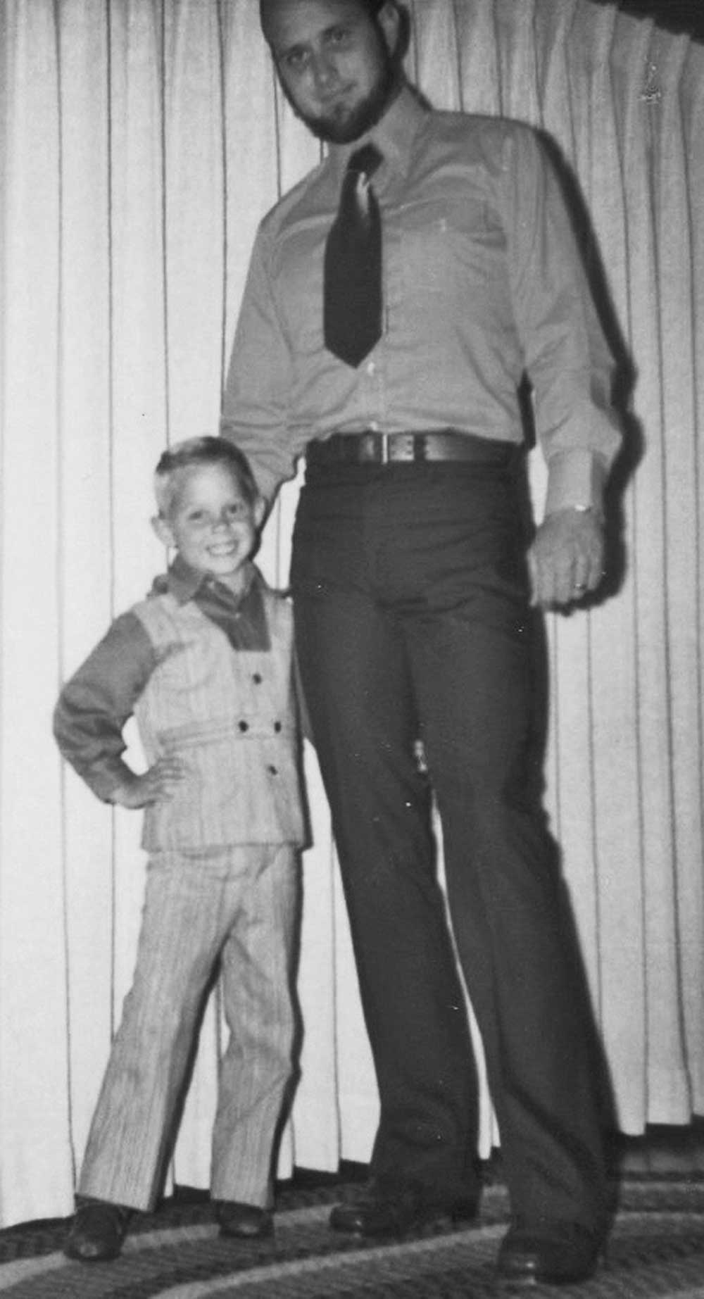 Old black and white photo of Brian as a little boy wearing a dress shirt and pants, and standing next to his dad, who is wearing pants with a button-up shirt and tie.
