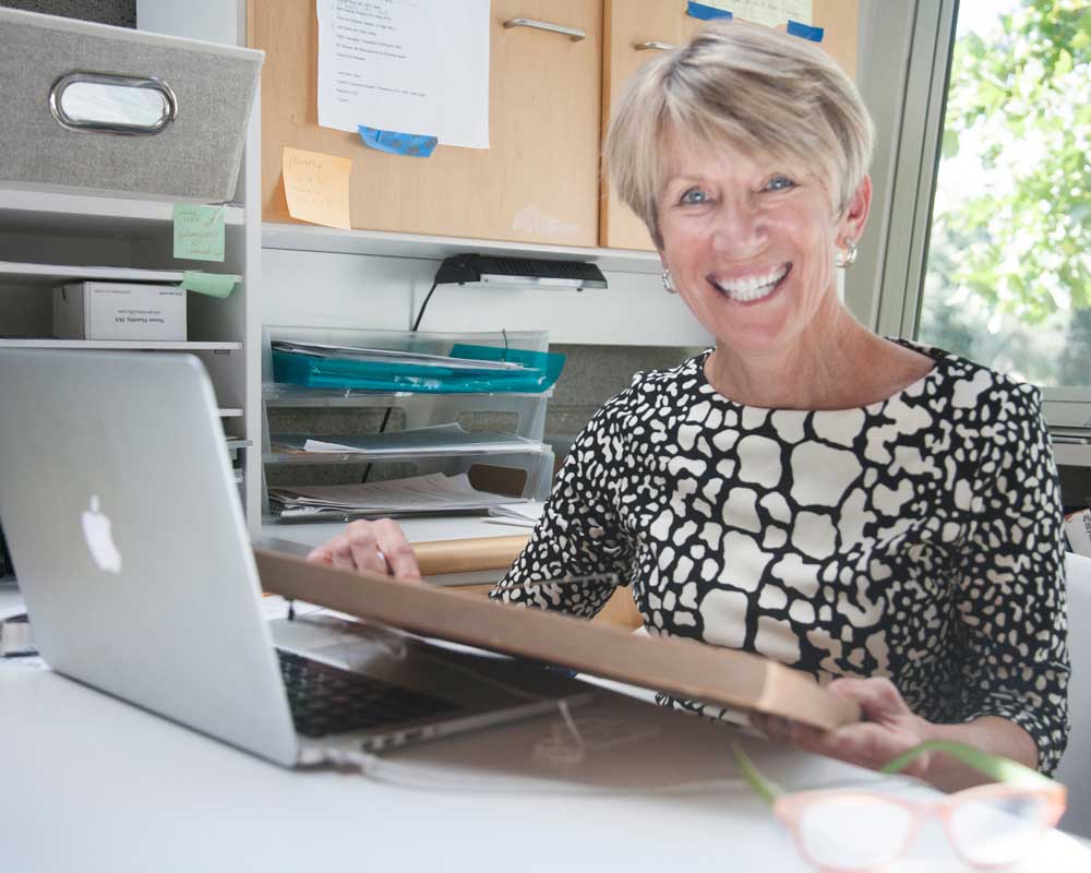 Close-up of Susan, a Witherell's employee, smiling at the camera while sitting at her desk. She has her laptop open in front of her and she is holding an auction item in her hands.