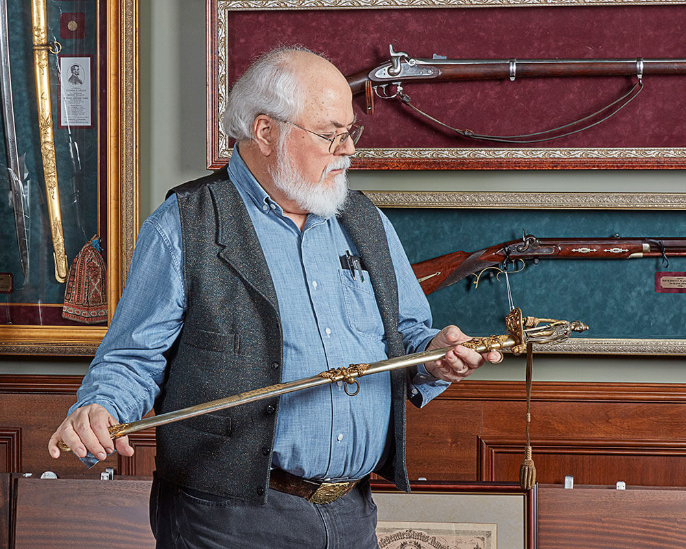Steven Crain, an older gentleman with a white beard and white hair, holds and examines the hilt of a sword while standing in front of other antique firearms and swords.