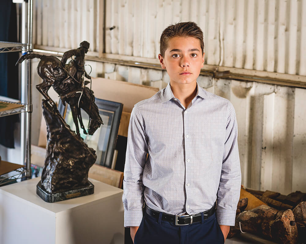 A young boy, Bennet Witherell, facing the camera and standing with hands in his pants pockets next to various auction items. He is wearing dark blue trousers, a dark belt, and a long-sleeved button-up shirt.