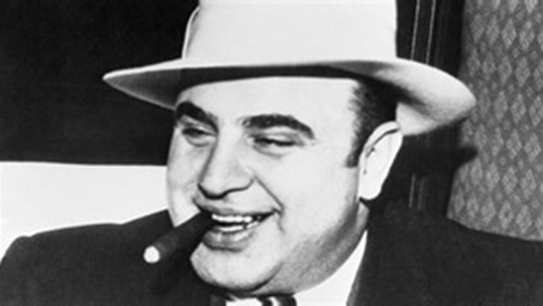 Close-up black and white photo of Al Capone's face. He is smiling with a cigar in his mouth and wearing a dark suit and light-colored hat.
