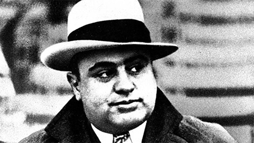 Black and white close-up of Al Capone's face. He is wearing a light-colored fedora with a black band around it, and he is looking off to his left. 