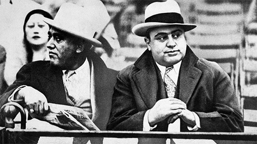 Black and white image of Al Capone and another man sitting next to each other. Both are wearing hats, dressed in suits and ties with heavy overcoats.