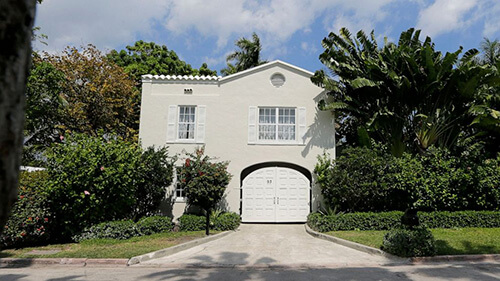 The gate house entrance of the wanterfont mansion once owned by Al Capone. It is a two-story white building with a flat front façade. There are two sets of windows on the second floor and one set on the first floor, wth the white gate to the right of it. The driveway is surrounded by green grass, shrubs, and trees on either side.