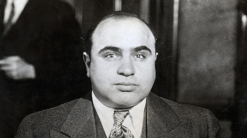 Close-up of Al Capone's face in a black and white image. He is wearing a dark suite and tie and looking at the camera, not smiling.