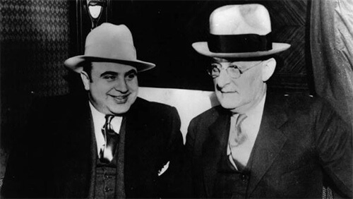 Close-up black and white photo of Al Capone smiling at an older gentleman sitting to his left. They are both wearing dark suits and ties with ligh-colored fedoras.