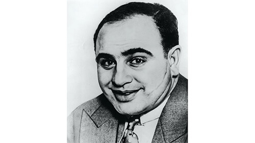 Close-up of a black and white headshot of Al Capone. He is smiling towards the camera, and wearing a suit and tie.