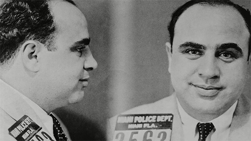 A mugshot of Al Capone, featuring his right-side profile and front profile. He is grinning slightly and wearing a light-colored suit and dark tie.