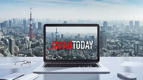 A screenshot of the title screen for a media show. It is a laptop sitting opened on a desk, and the words, 'Japan Today' showing on the screen. In the background is the view from a tall building of the city and buildings.