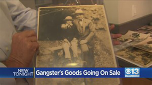 Screenshot from a news segment featuring a close-up of someone holding an old black and white photograph of Al Capone and his young son. The caption in the screenshot says,'Ganster's Goods Going on Sale'.