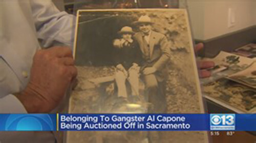 Screenshot from a news program featuring a close-up of a person holding up a black and white photo of Al Capone and his young son. The caption in the screenshot says, 'Belonging to Gangster Al Capone Being Auctioned Off in Sacramento'.