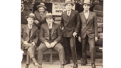 Old black and white group shot of Al Capone sitting on a bench alongisde four other men. All are dressed in suits with ties and wearing fedoras.