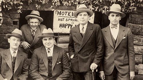 Old black and white group shot of Al Capone sitting on a bench alongisde four other men. All are dressed in suits with ties and wearing fedoras.