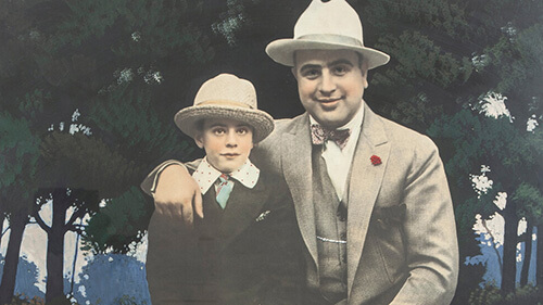 Close-up of a colorized version of Al Capone with his young son. They are both wearing suits with ties and fedoras.